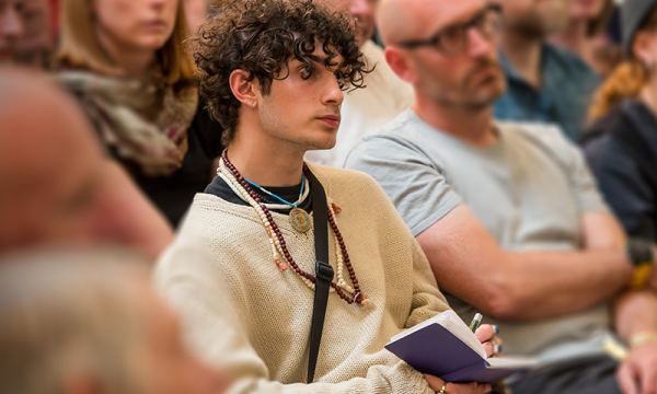 A young man with curly hair attentively listens while taking notes in a notebook during a lecture or discussion, surrounded by other engaged audience members.