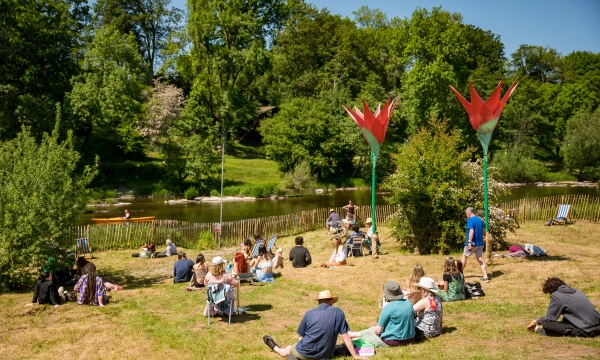 A sunny riverbank scene with people relaxing on the grass, some sitting, some drawing, near large flower-shaped sculptures and lush greenery.