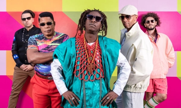A vibrant group of five musicians poses confidently against a colorful geometric backdrop, showcasing eclectic styles and accessories, highlighting their unique personalities and artistic flair.