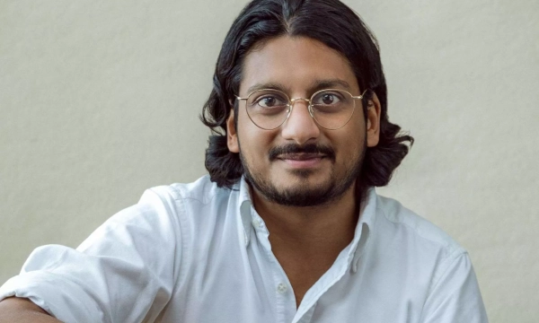 A smiling man with long, wavy hair and glasses, wearing a white shirt, poses in front of a simple, neutral background.
