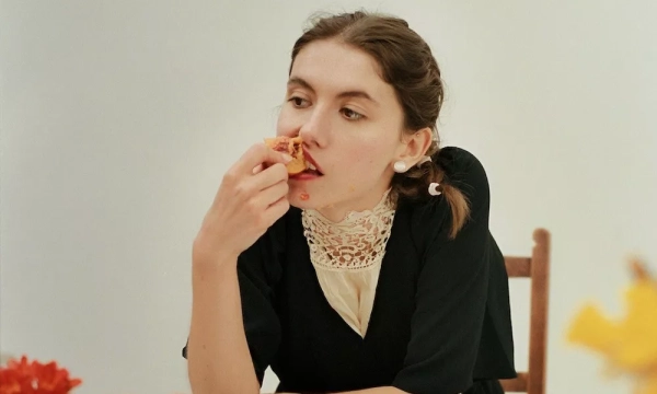 A young woman in a black dress with a lace collar sitting at a table, taking a bite of pizza, with a contemplative expression.