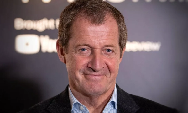 A middle-aged man with short, graying hair and a slight smile, wearing a dark jacket over a light blue shirt, set against a blurred background.