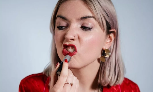 A young woman with blonde hair applies red lipstick, making a playful expression, while wearing a red patterned shirt and statement earrings.