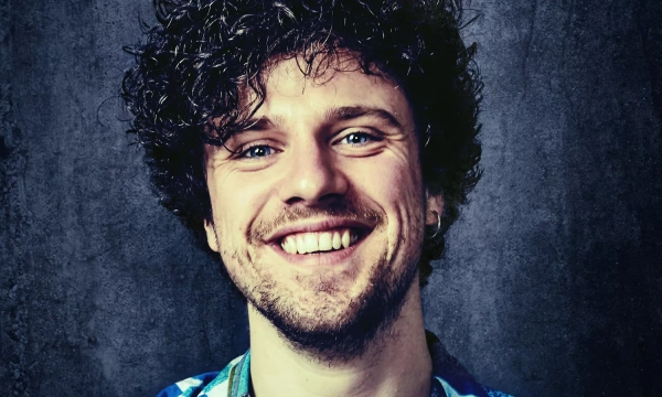 Smiling young man with curly hair wearing a colorful shirt, against a textured dark background.