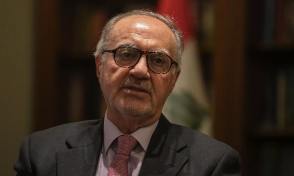 A man in a suit and glasses speaks, with a blurred background that includes a Lebanese flag and bookshelves.