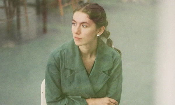 A young woman with a braided hairstyle sits thoughtfully in a casual setting, wearing a green coat and looking off to the side.