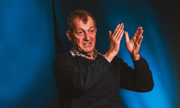 A middle-aged man gestures emphatically while speaking, set against a blue background, conveying a sense of urgency or strong emotion.