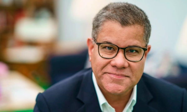 A middle-aged man with glasses smiles at the camera, wearing a dark suit jacket and light shirt, with a blurred background of a cozy, well-lit room.
