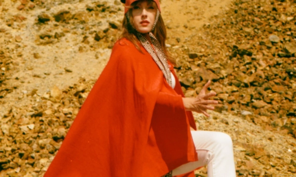 A model poses dramatically on rocky terrain, wearing a striking red cape, white pants, and tall red heels, with an artistic hat that resembles a totem.