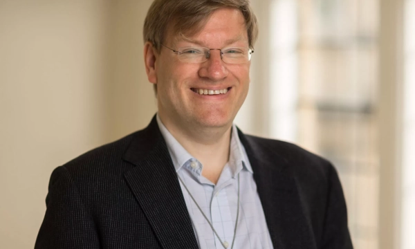 A smiling man with glasses and a pendant necklace, dressed in a black blazer over a light blue shirt, stands in a well-lit indoor setting.