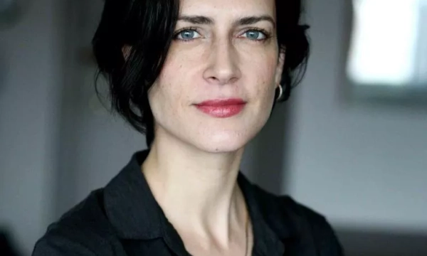 A woman with short black hair and striking blue eyes, wearing a black shirt, poses confidently against a softly blurred background.