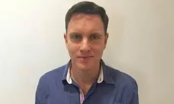 A man with short dark hair wearing a blue shirt stands against a plain background, smiling softly at the camera.