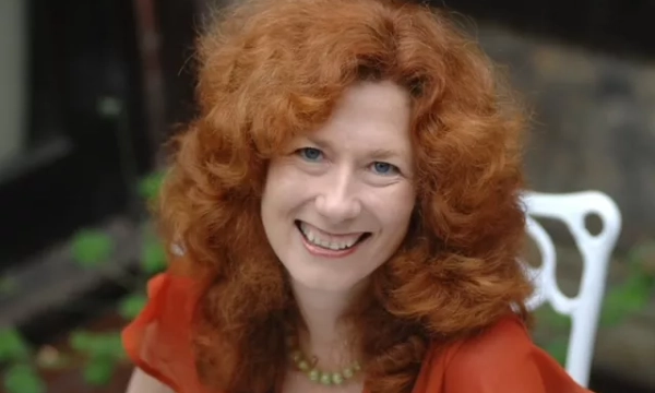 A smiling woman with curly red hair and an orange blouse, seated outdoors, surrounded by greenery.
