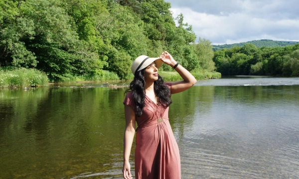 A woman in a pink dress and a straw hat stands by a calm river, surrounded by lush greenery and a partly cloudy sky.