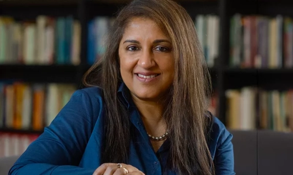 A smiling woman with long hair wearing a blue shirt, seated in front of a bookshelf filled with books.