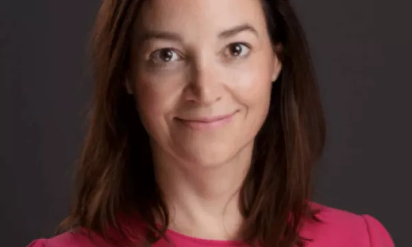 A woman with brown hair, wearing a pink blouse, smiles against a dark background, conveying a confident and professional demeanor.