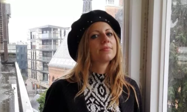 A woman with long hair wearing a black beret and patterned top poses by a window with a cityscape visible outside.