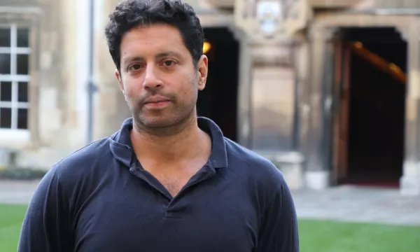 A man with dark, curly hair stands in front of an old building, casually dressed in a black polo shirt, with doors and window features visible behind him.