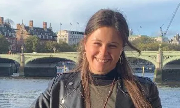 A smiling individual poses in front of a river with a historic bridge and ornate buildings in the background.