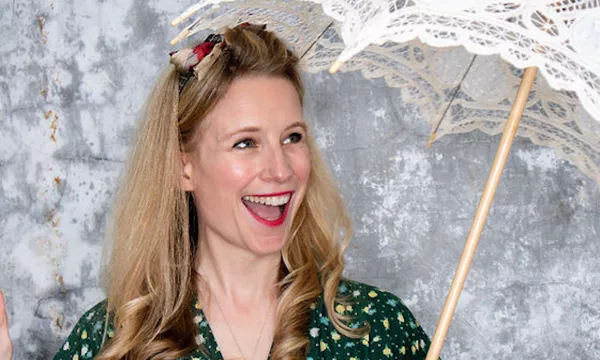 A woman with long, wavy hair and a playful expression holds a decorative lace parasol, wearing a green patterned dress.