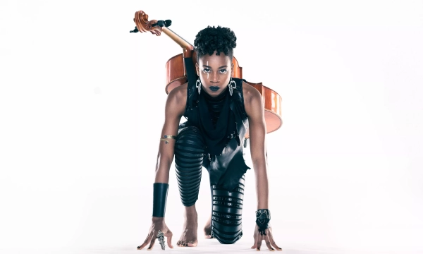 A person with a dramatic pose, dressed in leather attire, holding a microphone and a cello, positioned as if ready to perform.