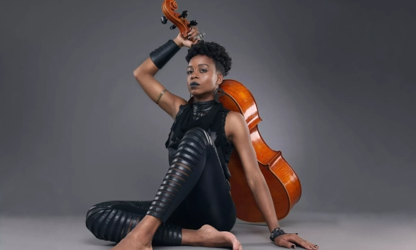 A stylish musician poses with a cello, showcasing a blend of modern fashion and artistic expression, featuring bare feet and striking accessories.