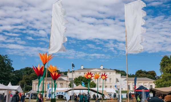 A vibrant outdoor festival scene featuring large floral decorations, white flags, and attendees mingling in front of a historic building under a partly cloudy sky.