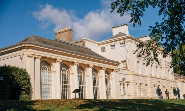 A historic building with classical architecture, featuring large windows and ornate columns, surrounded by trees and a clear blue sky.