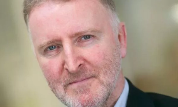 A close-up portrait of a middle-aged man with light hair and a beard, dressed in a suit, looking thoughtfully at the camera.
