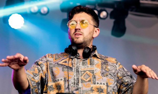 A DJ performs on stage, wearing yellow sunglasses and a patterned shirt, with headphones around his neck, while colorful lights illuminate the background.