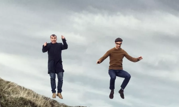 Two men joyfully jumping in mid-air against a cloudy sky, with a grassy landscape below them.