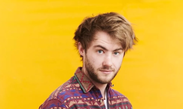 A young man with messy hair and a patterned shirt stands against a bright yellow background, looking directly at the camera.