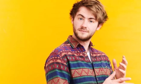 A young man with tousled hair wearing a colorful patterned shirt poses against a bright yellow background, gesturing with his hands.
