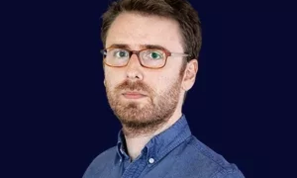 A man with glasses and a beard, wearing a blue button-up shirt, poses against a dark background.