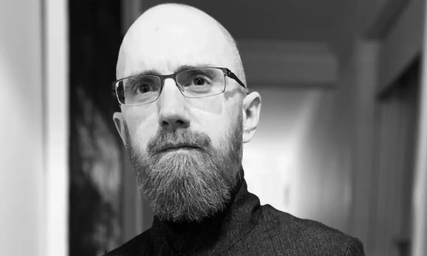 A man with a bald head and a beard, wearing glasses and a dark turtleneck, gazes thoughtfully with a blurred hallway in the background.