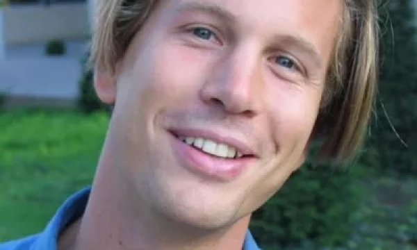 A smiling young man with blond hair, wearing a blue shirt, stands outdoors in a green setting.