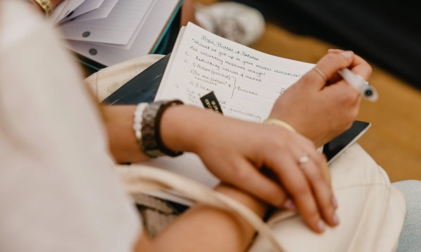 A person's hands resting on a notebook filled with handwritten notes, featuring a pen and loose papers in the background.