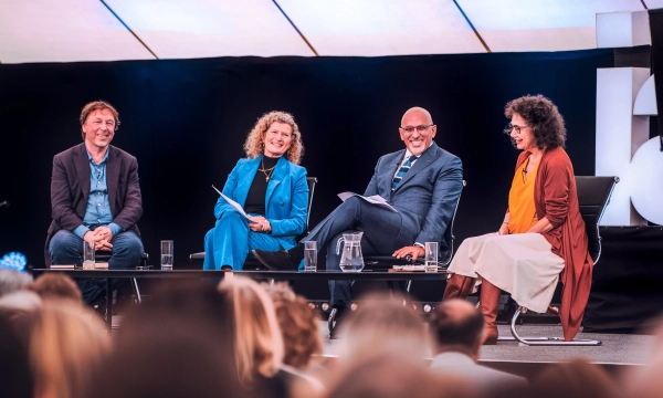 A panel discussion featuring four speakers seated at a table, engaged in conversation, with a modern backdrop and audience in soft focus.