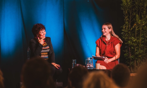 Two women sit on stage during an event, engaging in conversation. One woman has curly hair and wears a black jacket, while the other, in a red dress, holds notes and smiles.