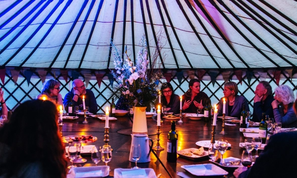 A group of people seated at a large round table inside a yurt, enjoying a candlelit dinner with floral arrangements and various dishes.