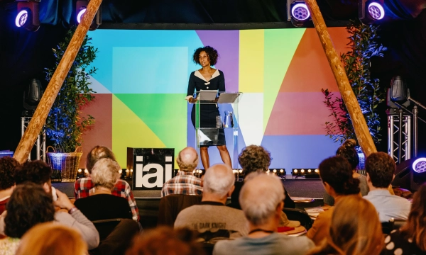 A speaker stands at a podium in front of a colorful backdrop, addressing an audience during an event at an indoor venue.