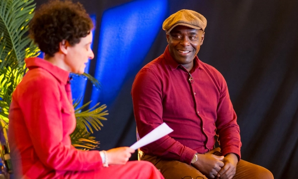 A man in a brown shirt and cap smiles while seated across from a woman in a red dress during an interview or discussion.