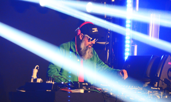 A DJ with a white beard and sunglasses performs on stage, surrounded by colorful light beams and electronic equipment.