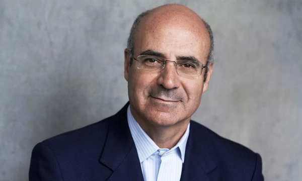 A middle-aged man with a bald head and glasses, dressed in a dark suit, smiles against a neutral background.