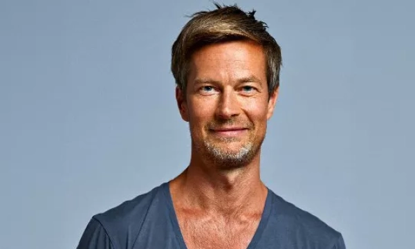 A smiling man with short, tousled hair and a light beard, wearing a gray V-neck shirt against a plain background.