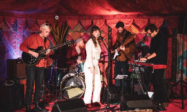 A band performing on stage in a colorful, decorated environment, with a singer in a white outfit and four musicians playing various instruments.