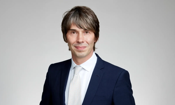 A well-dressed man with medium-length hair, smiling at the camera, against a plain background. He wears a dark suit and a light-colored tie.