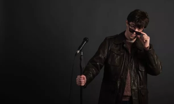A young man in a leather jacket adjusts his sunglasses while standing next to a microphone against a dark background.