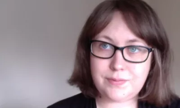A woman with shoulder-length brown hair and glasses, wearing a black top, is looking slightly to the side against a neutral background.