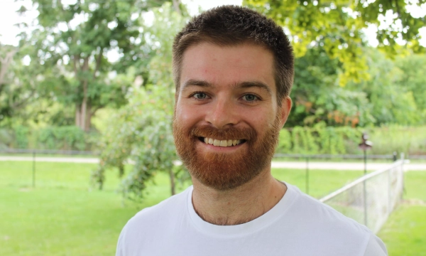 A smiling man with a reddish beard and short hair stands outdoors in a green park setting.